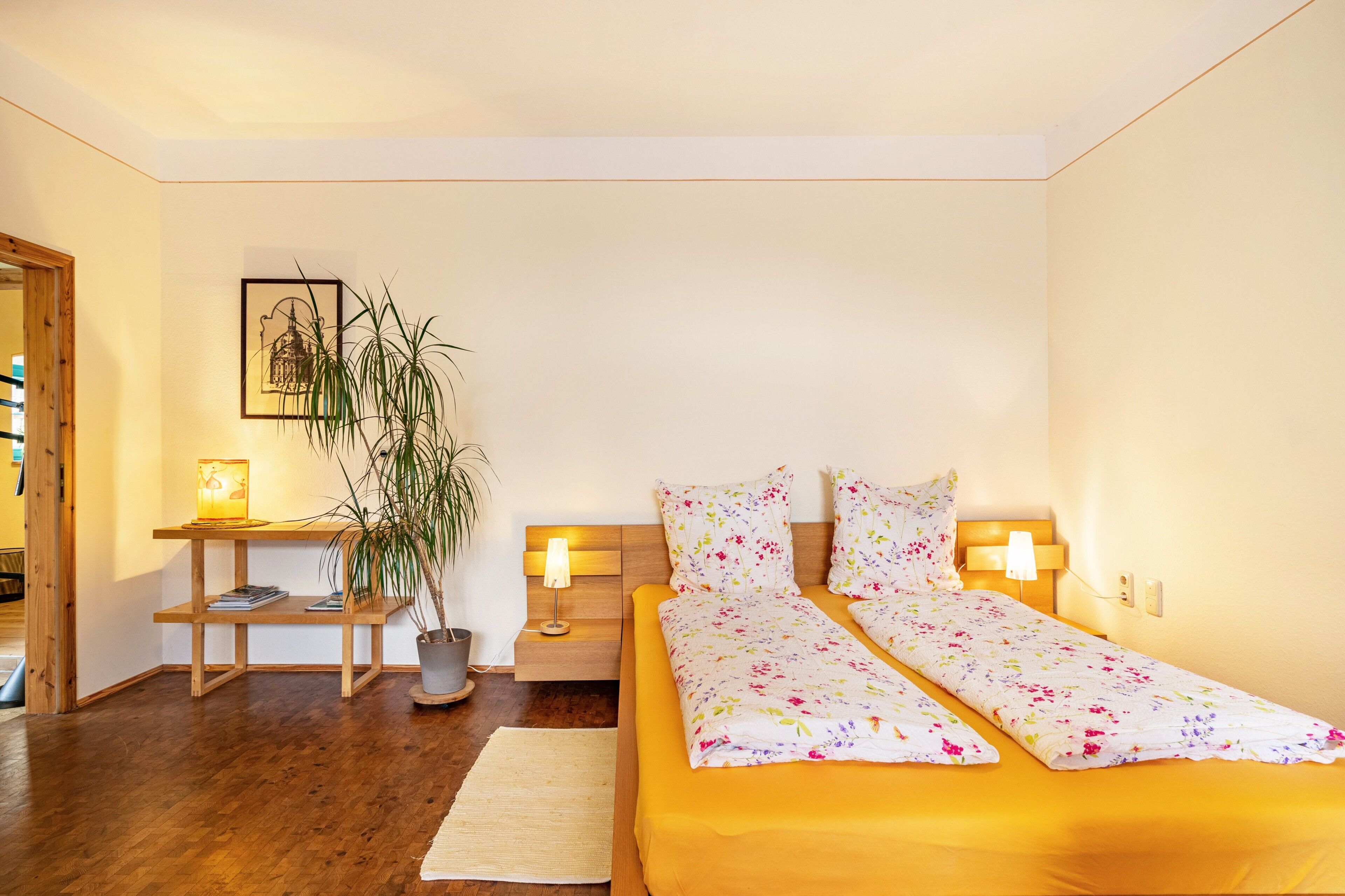 Un dormitorio con cama, mesitas de noche, lámpara, planta y un cuadro en la pared.