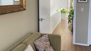 A living room with a green sofa, a patterned pillow, a large mirror, and a door leading to a hallway with plants. - 3-bedroom house in awesome East Hampton with AC, WiFi, backs up to county park (East Hampton)