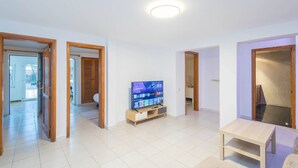 Un amplio salón con una televisión de pantalla plana sobre un soporte de madera, piso de baldosas claras y puertas de madera que dan a otras habitaciones.