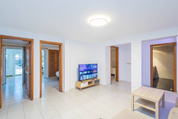 Un amplio salón con una televisión de pantalla plana sobre un soporte de madera, piso de baldosas claras y puertas de madera que dan a otras habitaciones.