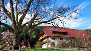 Ein zweistöckiges Gebäude mit rotem Ziegeldach, ein großer Baum im Vordergrund und ein Spielplatz mit Schaukeln und Rutschen.