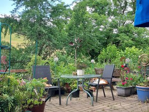 En udendørs terrasse med et bord og stole, omgivet af krukker med planter og en have.