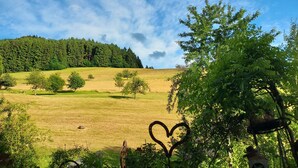 A garden with a heart-shaped metal sculpture, a table with chairs, and a view of a green field and forest under a partly cloudy sky.