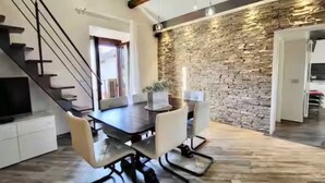 Una cocina moderna con un muro decorativo de piedra, un comedor con mesa de madera oscura y sillas blancas, y una escalera con barandilla negra.