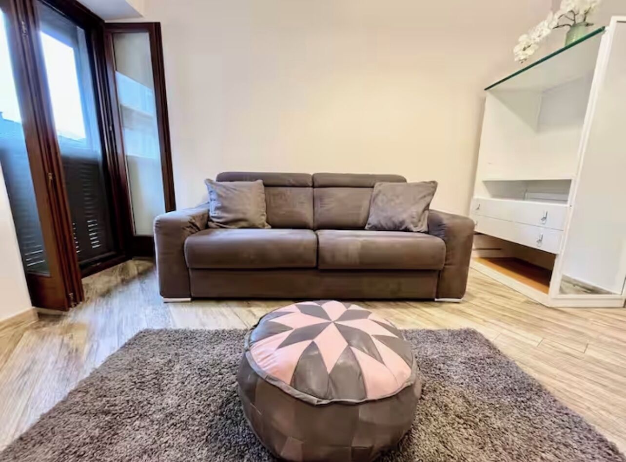 A living room with a brown sofa, a patterned ottoman, and a white shelving unit.