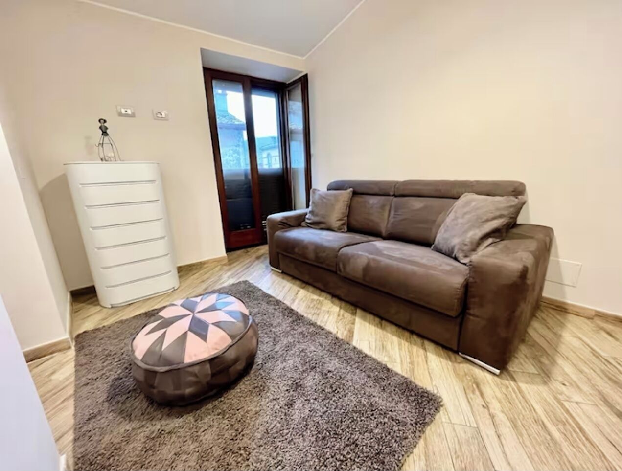 A living room with a brown leather sofa, a patterned ottoman, a white cabinet, and a door leading outside.