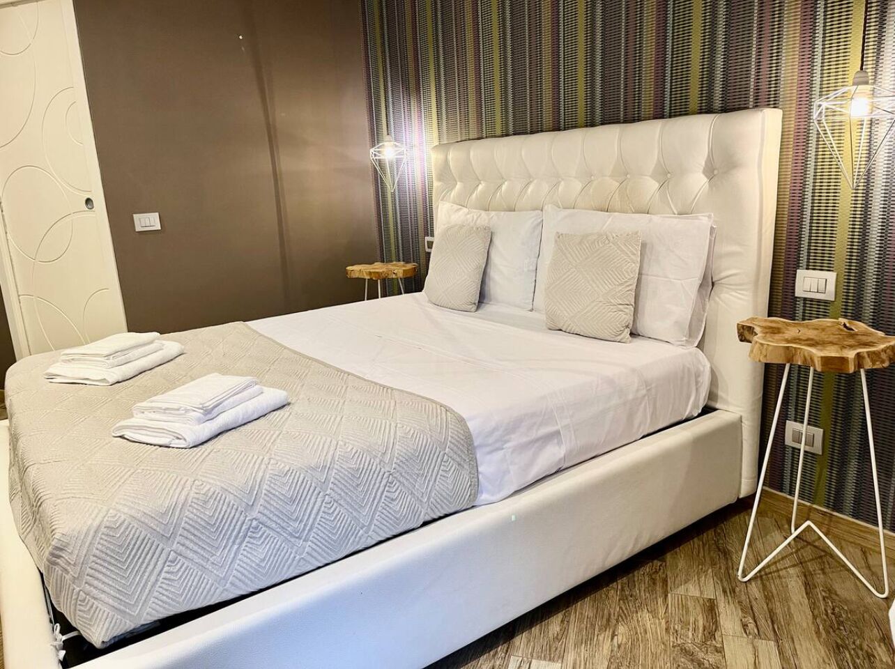 Un lit bien fait, avec une tête de lit blanche et des oreillers, une table de chevet en bois et un mur à rayures en arrière-plan.