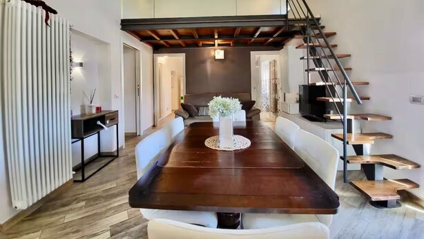 Un comedor moderno con una larga mesa de madera, sillas blancas acolchadas y un jarrón central. Una escalera de madera con barandilla metálica conduce a un nivel superior.
