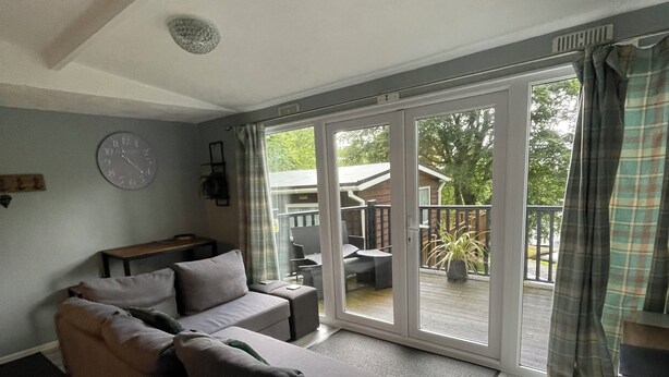A modern living room with a grey sectional sofa, a wooden floor, and a large sliding glass door leading to an outdoor patio area. - Delightful 2 Bedroom Welsh Chalet, situated between Snowdonia and Caernarfon. (Caernarfon)