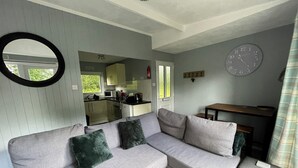 A modern living room with a grey sectional sofa, a round mirror, a wooden table, and a kitchen in the background. - Delightful 2 Bedroom Welsh Chalet, situated between Snowdonia and Caernarfon. (Caernarfon)