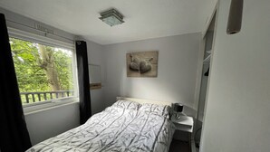 A bedroom with a bed, a bedside table, a lamp, a window with a view of trees, and a wall-mounted artwork. - Delightful 2 Bedroom Welsh Chalet, situated between Snowdonia and Caernarfon. (Caernarfon)