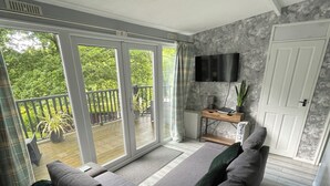 A modern living room with a large sliding glass door leading to a balcony, a flat-screen TV mounted on the wall, and a cozy seating area with a sofa and cushions. - Delightful 2 Bedroom Welsh Chalet, situated between Snowdonia and Caernarfon. (Caernarfon)