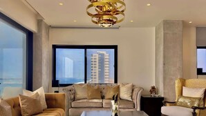 A modern living room with a sofa, armchair, and coffee table, a large window with a city view, and a decorative chandelier.