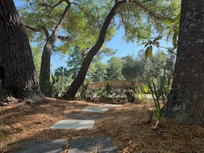 A stone pathway leading through a wooded area with large trees and a wooden bridge in the background.