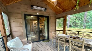 Une terrasse en bois avec une table et des chaises, un fauteuil en osier avec un coussin, et une porte vitrée donnant sur un espace intérieur.
