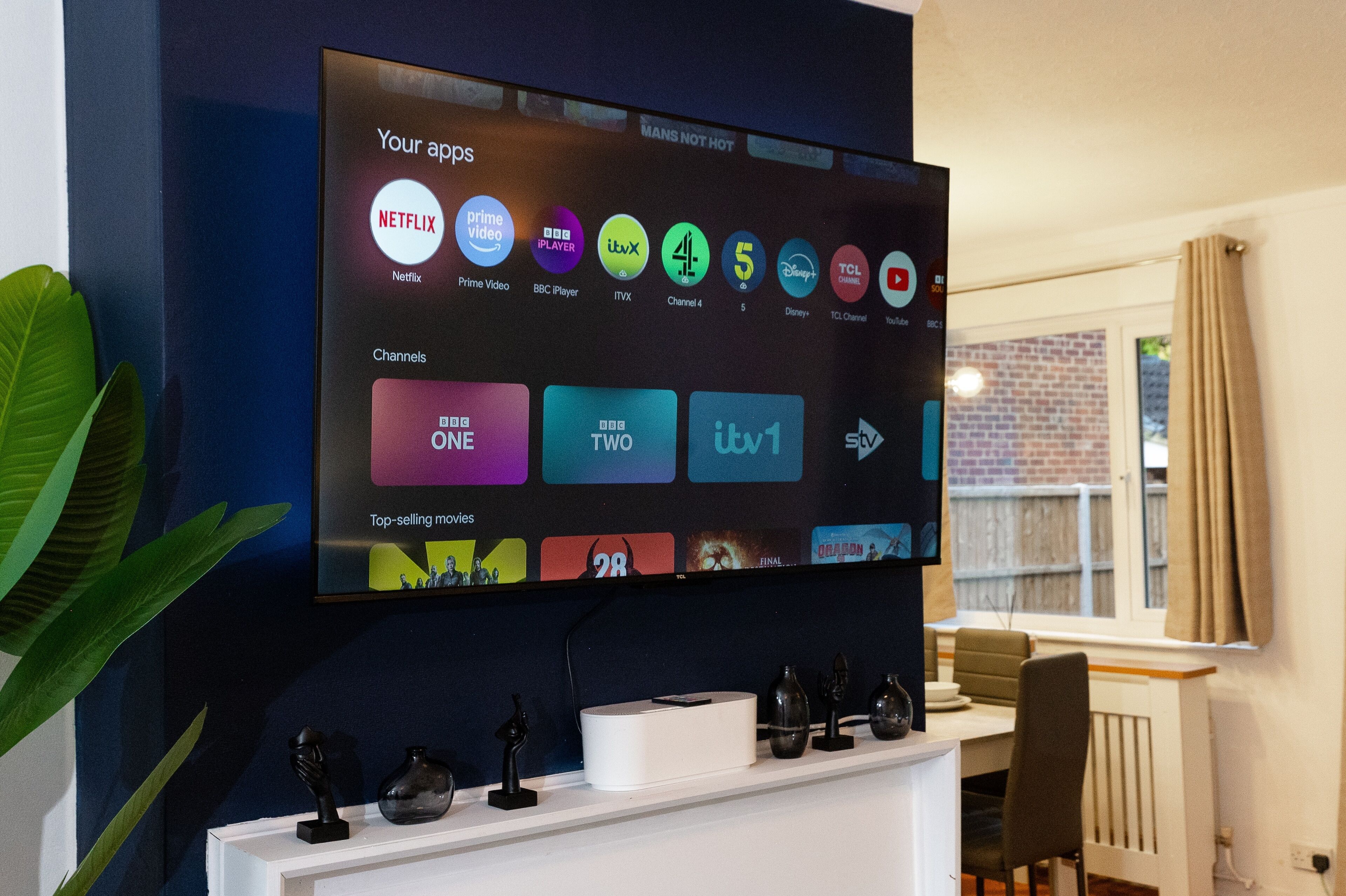 A flat-screen TV mounted on a wall displaying a streaming service interface with various channel logos and a list of top-selling movies.