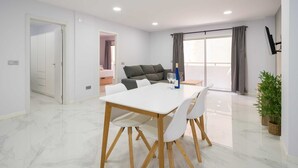 A modern living room with a white dining table, grey sofa, and a large window with curtains. - Holiday Home Near Playa de Las Canteras (Las Palmas de Gran Canaria)