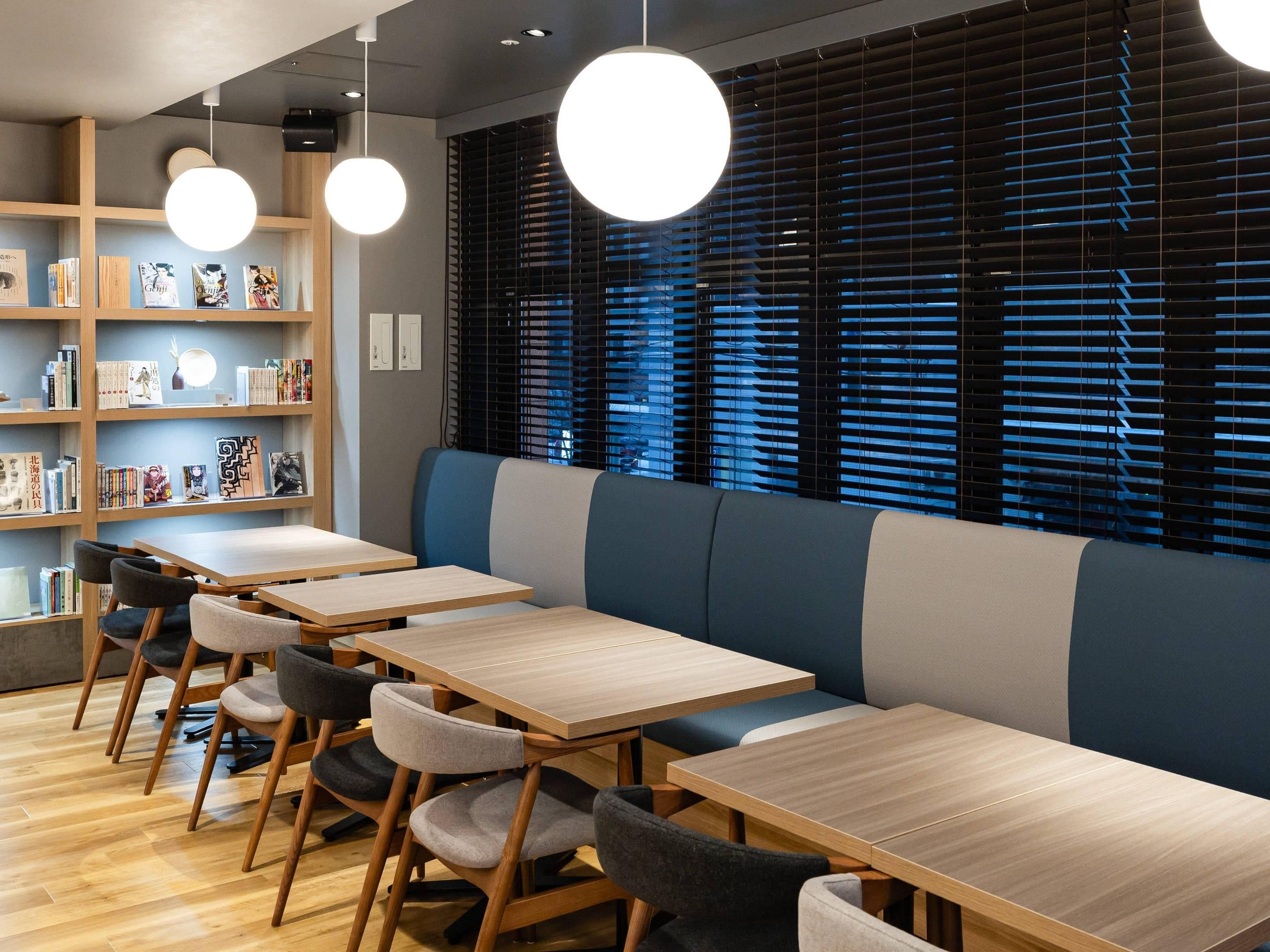 A modern library or reading area with wooden tables and chairs, bookshelves, and large windows with blinds.