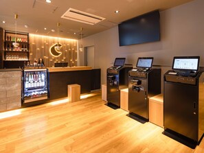 Un espace bar moderne doté d’un comptoir en bois, d’un présentoir réfrigéré pour les boissons et de bornes de commande en libre-service.