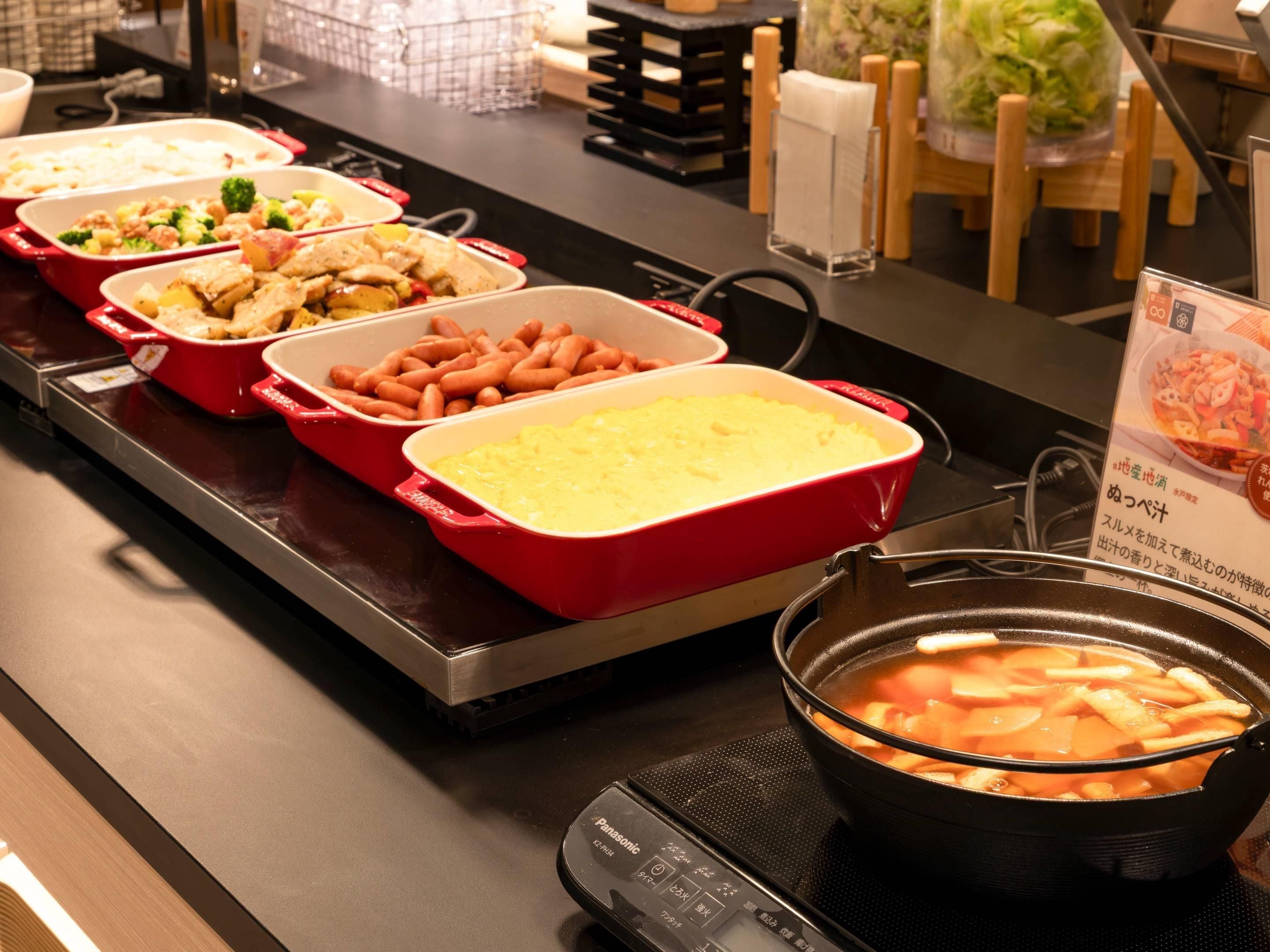 A buffet table with various dishes in red and white containers, a black pot with a soup, and a Panasonic electric stove.