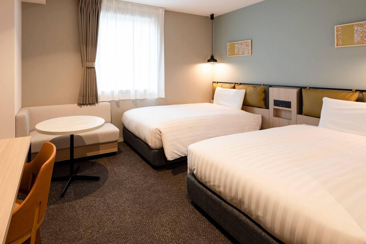 A hotel room with two beds, a round table, a sofa, and a window with curtains.