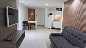Un salon moderne avec un canapé gris, un téléviseur fixé sur un mur en bois, une petite table avec deux chaises et, au fond, un coin cuisine.