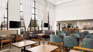 Un hall de hotel espacioso con varias zonas de estar, incluyendo sofás, sillas y mesas. Se observan amplias ventanas con cortinas, un televisor montado en la pared y una cocina al fondo.