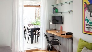 Une chambre moderne avec un bureau en bois, un ordinateur et une télévision. On aperçoit, à travers une fenêtre, un coin repas avec des chaises et une table.