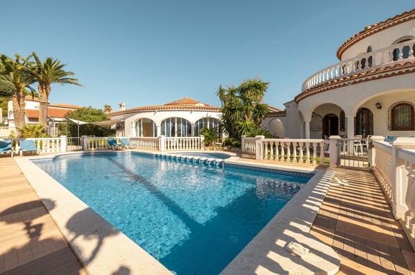 A swimming pool with a tiled deck, palm trees, and a building with arched windows and a tiled roof.