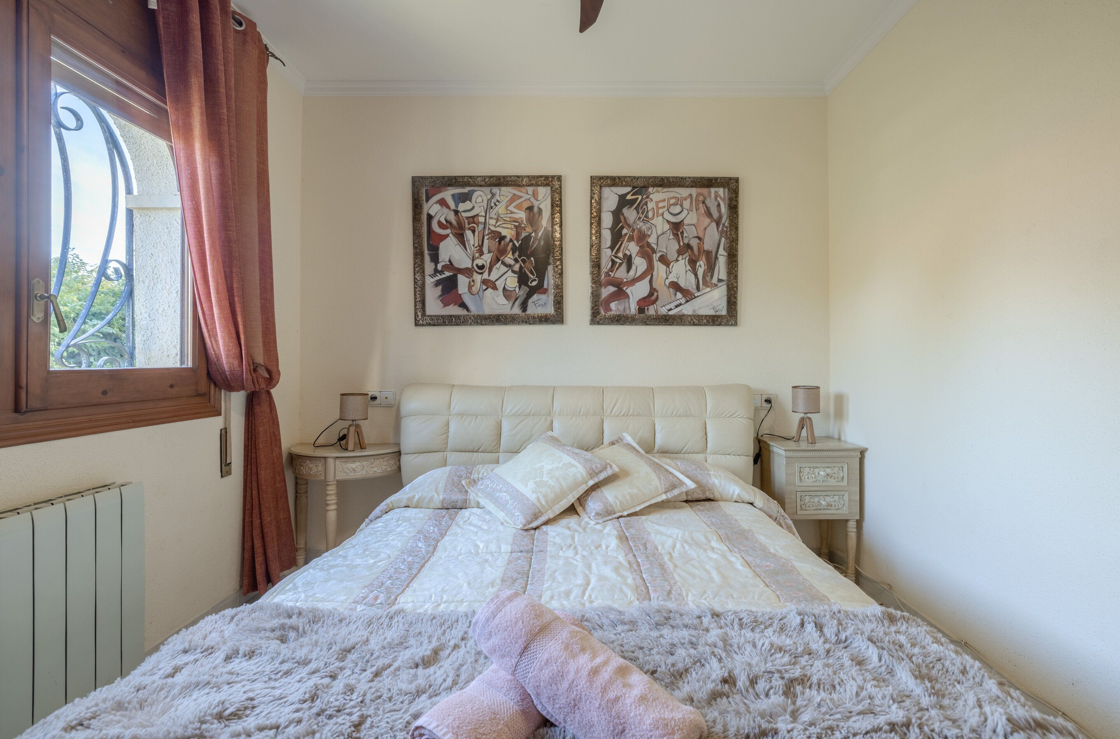 A bedroom with a bed, two wall-mounted paintings, a window with curtains, bedside tables with lamps, and a radiator.