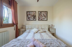 A bedroom with a bed, two wall-mounted paintings, a window with curtains, bedside tables with lamps, and a radiator.