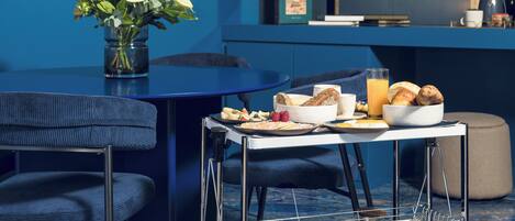 A modern room with a blue wall, a dining table with a tray of food, a chair, and a small cart with snacks.