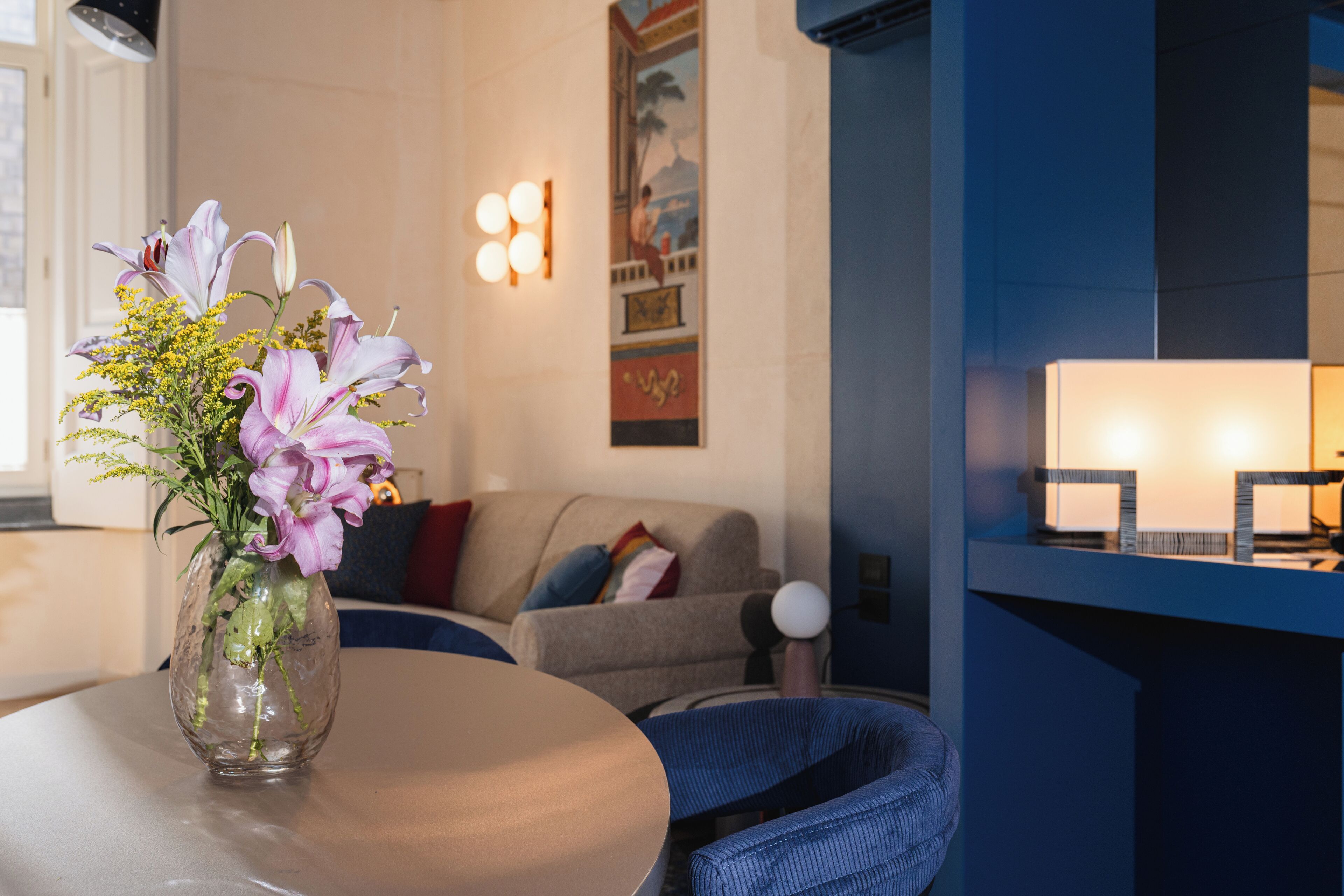 A modern living room with a round table, a vase of flowers, a sofa, and a wall-mounted lamp.