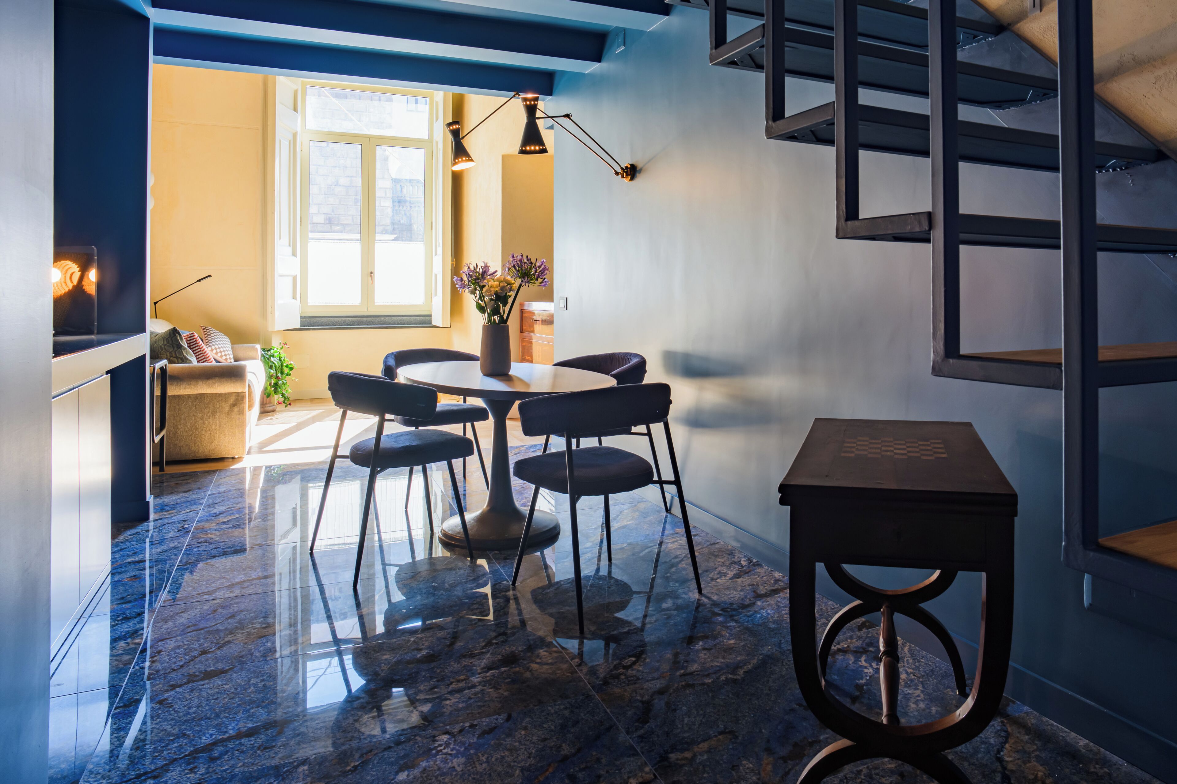 A modern interior with a marble floor, a round table with chairs, a small wooden stool, and a staircase.