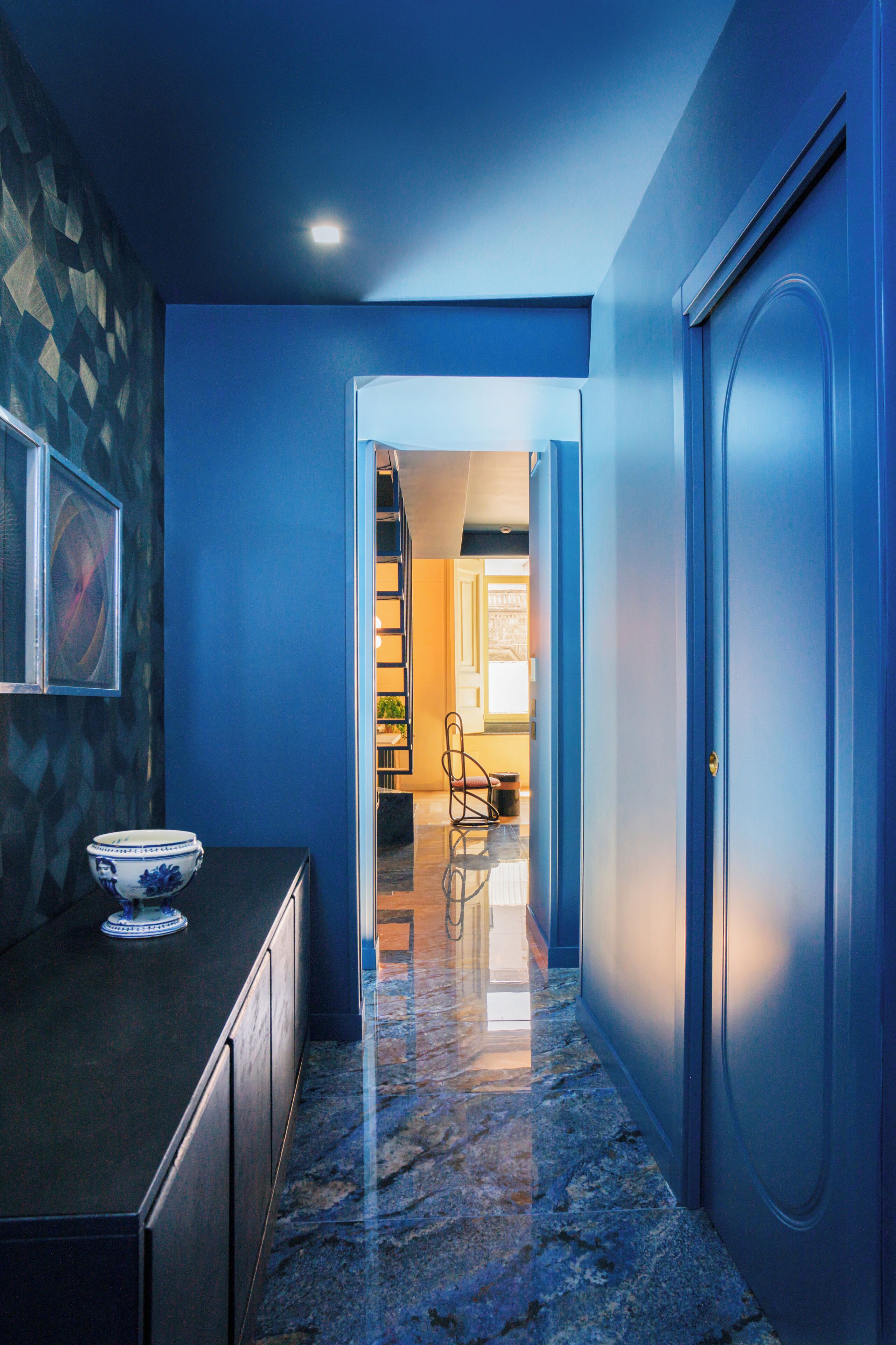Un pasillo con pared azul, suelo de mármol y una puerta que da a otra habitación.