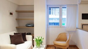 A compact living space with a white sofa, a small table with a remote control, a beige armchair, a window with a view of a balcony, and a small shelf with a speaker.