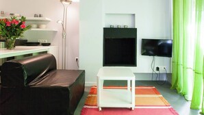 A modern living room with a black leather sofa, a white side table, a floor lamp, a flat-screen TV mounted on the wall, and a green curtain.