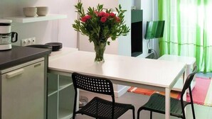 A modern kitchen with a white countertop, a coffee maker, and a vase with red flowers. There are two black chairs and a green curtain in the background.