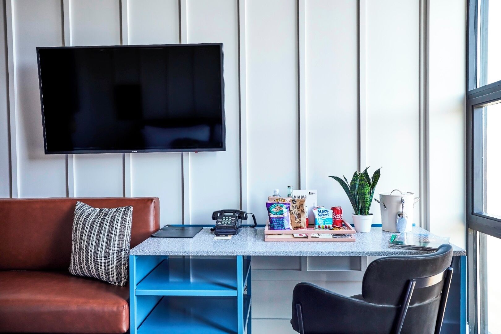 Un salon moderne avec un canapé en cuir marron, une table basse sur laquelle sont posés des en-cas et un téléphone, un téléviseur fixé à un mur recouvert de panneaux blancs, et une plante en pot.