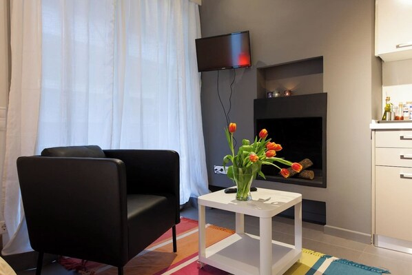 Ein modernes Wohnzimmer mit einem schwarzen Ledersofa, einem kleinen weißen Beistelltisch mit einer Tulpenvase, einem flachbildfernseher an der Wand und einem Kamin mit Kaminfeuer.