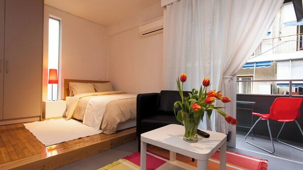 Una habitación de hotel moderna con una cama, una mesita con un jarrón de flores, una silla roja y un balcón con vistas.