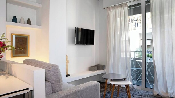 Un soggiorno moderno con un divano grigio, un televisore a schermo piatto montato a parete, un piccolo tavolino rotondo e un balcone con tende bianche.