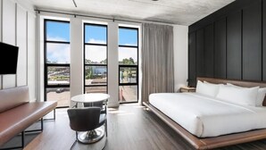 Une chambre d’hôtel moderne, dotée d’un grand lit, d’un bureau et d’une chaise. De grandes fenêtres offrent une vue sur l’extérieur.