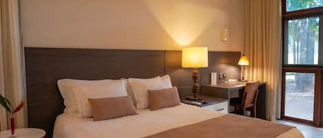 A hotel room with a bed, bedside lamps, a desk, and a view of trees through the window.