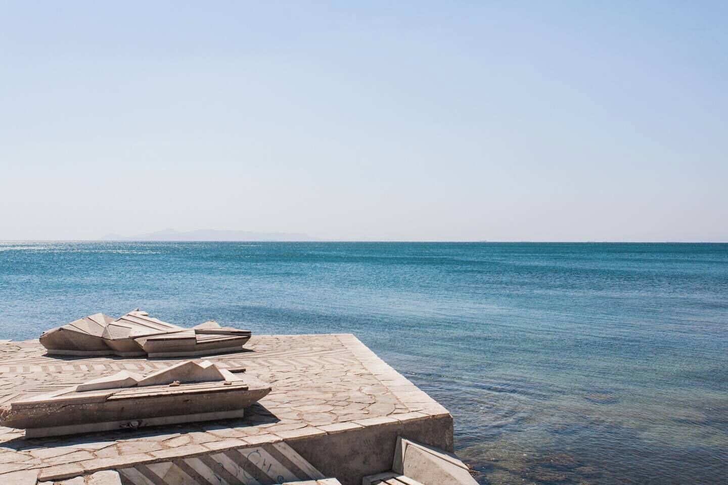 A stone pier extending into calm blue waters with clear skies.