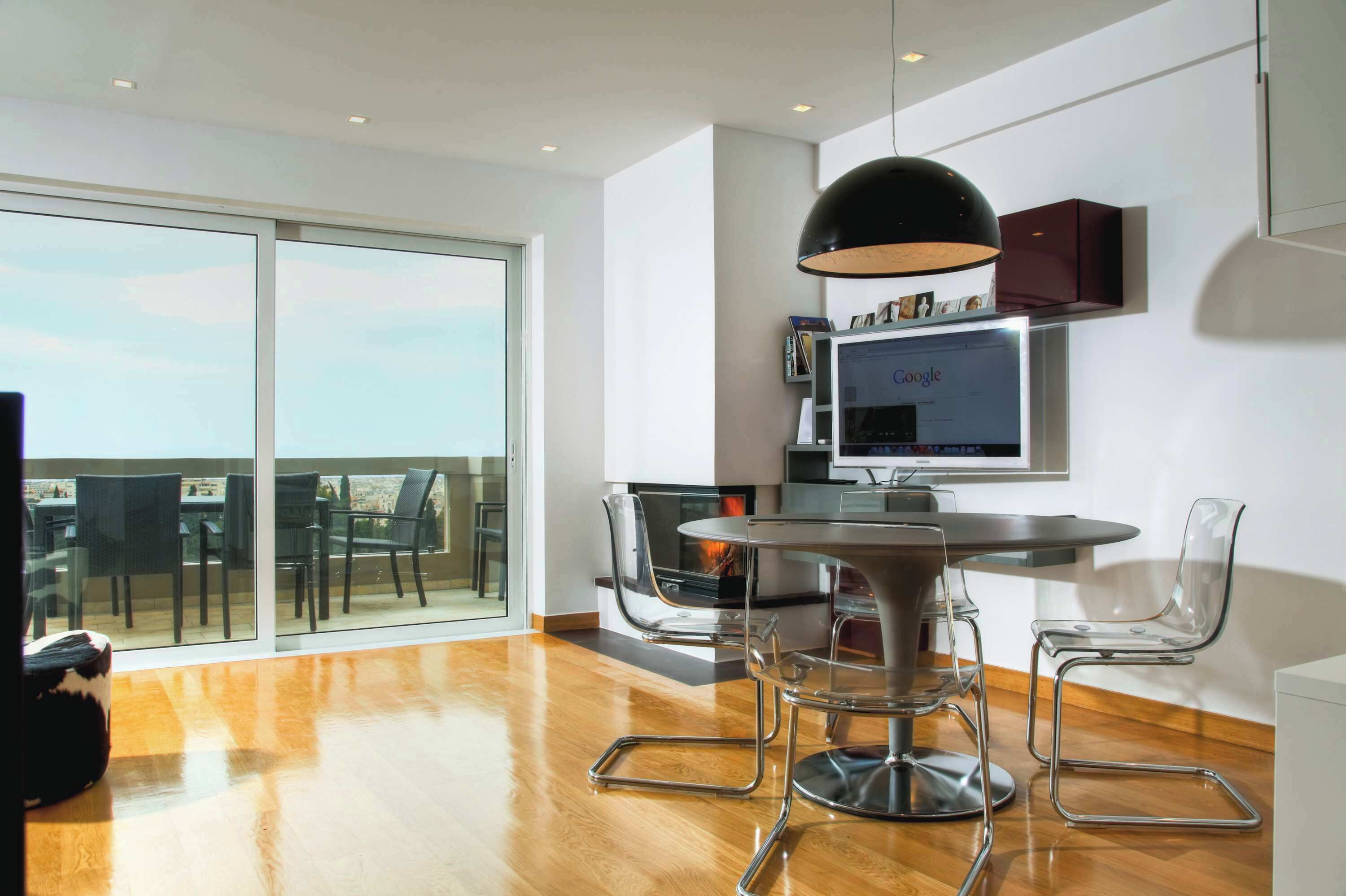 A modern dining area with a round table, clear chairs, a fireplace, and a large window offering a view.