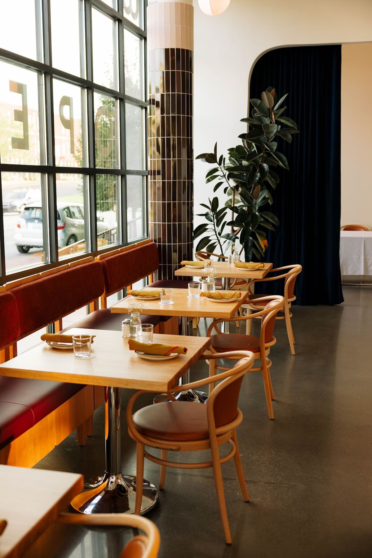 Un restaurante con mesas y sillas de madera, ventanas amplias y una planta en maceta.