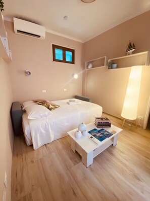 A bedroom with a bed, bedside table, bedside lamp, air conditioning unit, window, and a shelf with books.