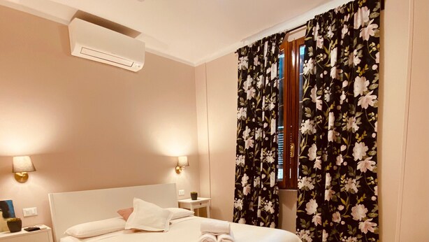 Un dormitorio ordenado con cama, lámparas de noche, mesita de noche, una silla y una ventana con cortinas florales.