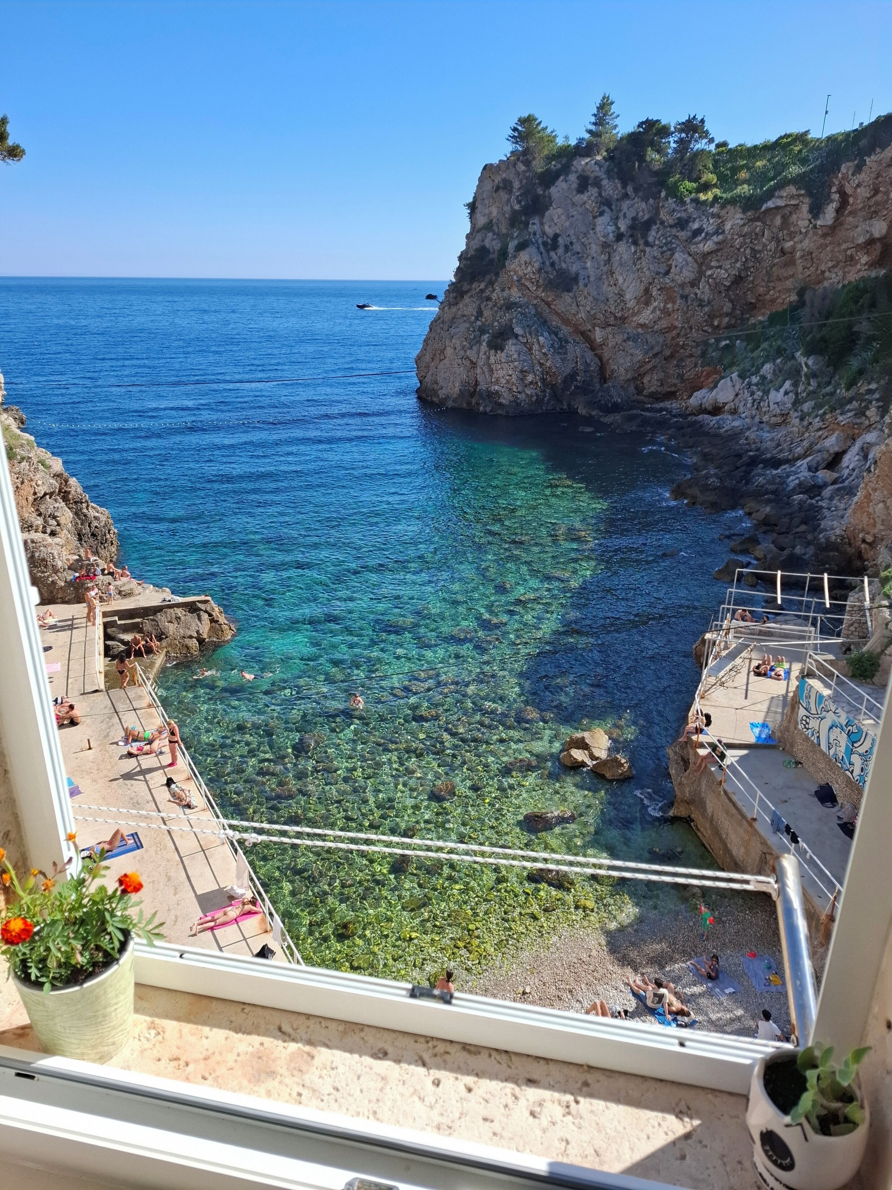 A coastal view from a balcony with clear water, rocky cliffs, and a few people.